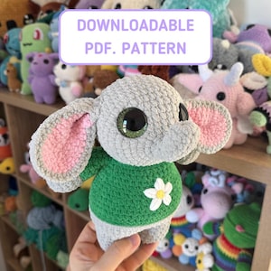 May include: A gray crocheted elephant with large, green eyes and pink-lined ears. It wears a green sweater with a white flower. The image includes the text "DOWNLOADABLE PDF. PATTERN".