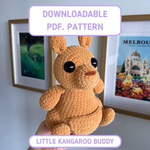 May include: A light brown crocheted kangaroo plush toy with large black eyes and a small brown nose. The text DOWNLOADABLE PDF. PATTERN and LITTLE KANGAROO BUDDY is displayed in white boxes with purple borders above and below the plush toy.
