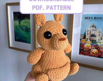 May include: A light brown crocheted kangaroo plush toy with large black eyes and a small brown nose. The text DOWNLOADABLE PDF. PATTERN and LITTLE KANGAROO BUDDY is displayed in white boxes with purple borders above and below the plush toy.