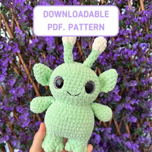 May include: A green crocheted alien plush toy with large black eyes and a white crocheted antenna. The toy is being held in a hand against a background of purple flowers.