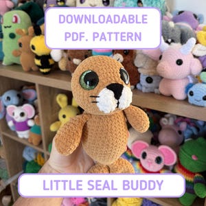 May include: A crocheted seal plushie with large, glittery green eyes and a black nose and whiskers. The seal is tan with white accents and is labeled "LITTLE SEAL BUDDY" and "DOWNLOADABLE PDF. PATTERN". Other crocheted toys are visible in the background.