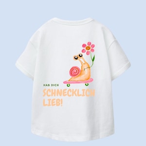 Könnte beinhalten: Weißes T-Shirt mit einem Cartoon-Schnecken-Design auf der Rückseite. Die Schnecke fährt Skateboard und hält eine Blume. Pfirsichfarbener Text lautet "HAB DICH SCHNECKLICH LIEB!". Schnecke und Blume sind orange und rosa.