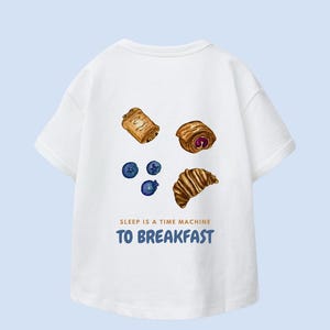 Könnte beinhalten: Weißes T-Shirt mit Aquarellillustrationen von Gebäck und Blaubeeren. Der Text "SLEEP IS A TIME MACHINE TO BREAKFAST" ist darunter in Blau gedruckt.