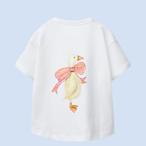 Könnte beinhalten: Weißes T-Shirt mit einer Aquarellillustration einer Ente mit rosa Schleife. Die Ente hat gelbe Federn, einen orangen Schnabel und orange Füße. Das Shirt hat kurze Ärmel und einen Rundhalsausschnitt.
