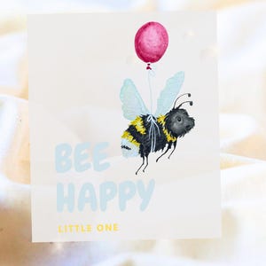 Könnte beinhalten: Aquarellillustration einer Biene, die einen rosa Ballon hält. Die Biene hat gelbe und schwarze Streifen und blaue Flügel. Die Worte "BEE HAPPY LITTLE ONE" sind in Hellblau und Gelb geschrieben.