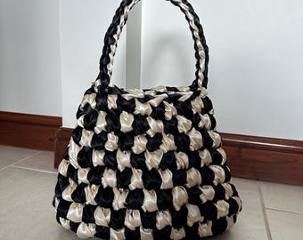 May include: A black and cream-colored handbag with a woven design. The bag features a rounded shape and a braided handle. The woven pattern creates a checkerboard effect with alternating black and cream-colored satin ribbons. The bag is sitting on a tiled floor.