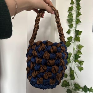 May include: A handmade handbag with a braided brown handle and a body made of interwoven blue and brown satin ribbons. The bag is held up by a hand against a white background with green vines.