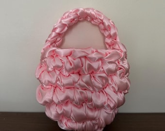 May include: A small, rounded handbag crafted from light pink satin ribbon. The body of the bag features a gathered, textured design, while the handle is constructed from a thick, braided satin ribbon in the same matching shade of light pink.