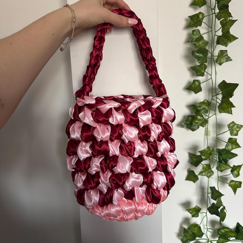 May include: A handmade, small tote bag with a woven design. The bag is made of alternating pink and burgundy satin ribbons. The handles are braided burgundy ribbons. The bag is held by a person.