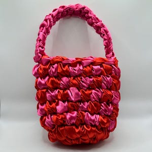 May include: A handmade, small tote bag in a vibrant combination of red and pink satin ribbons. The bag features a braided handle and a body constructed from interwoven ribbons, creating a textured, eye-catching design. The bag is suitable for carrying small items.