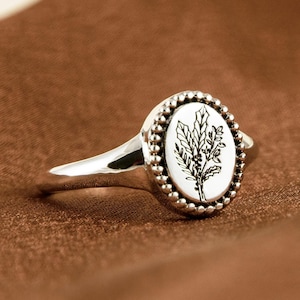 May include: A silver ring with an oval, white face featuring a black botanical design. The face is surrounded by a beaded border. The ring is set against a brown background.