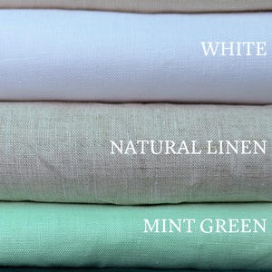 May include: A stack of five folded fabric swatches in beige, white, natural linen, mint green, and emerald green. Each color is labeled with its corresponding name in white text, clearly visible on the surface of each respective fabric layer.