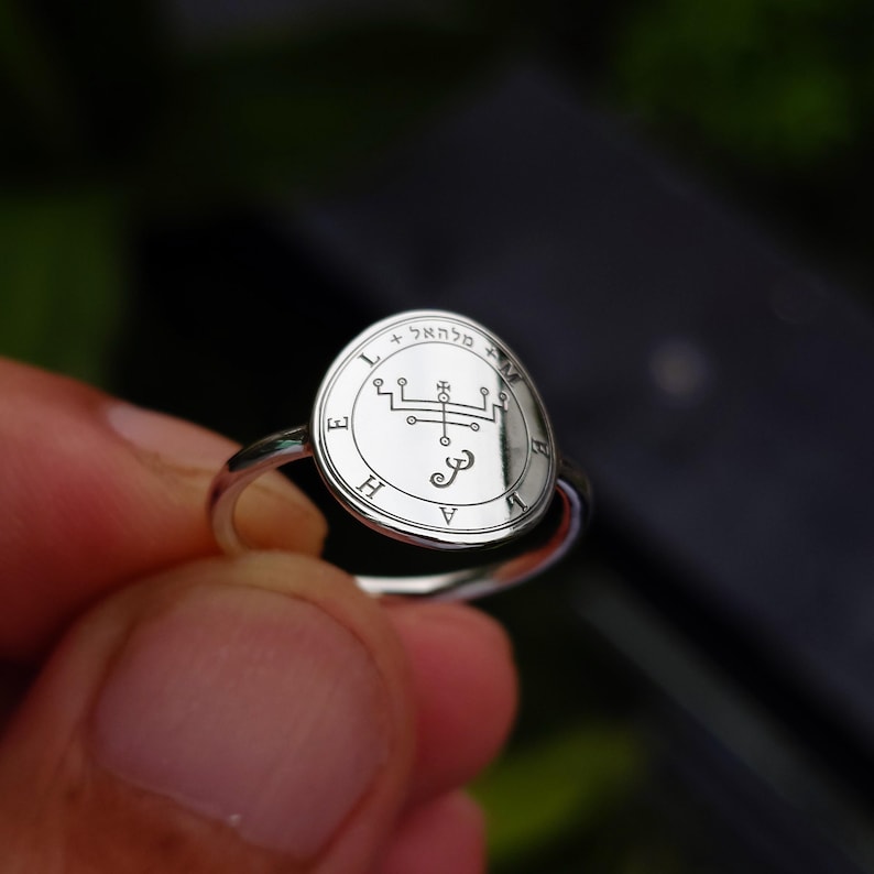 May include: A silver ring with a circular, engraved design. The ring features a detailed emblem with Hebrew text and symbols. The ring is held up against a blurred background, highlighting the intricate design and metallic sheen.