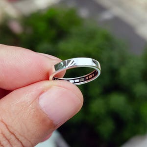 Peut inclure: Une bague argentée polie dotée d'une bande extérieure lisse et réfléchissante, avec une surface intérieure sombre ornée d'une série de petits accents métalliques rectangulaires et circulaires disposés en motif répétitif sur la circonférence.