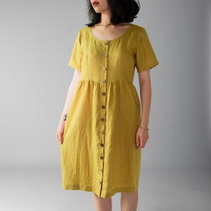 May include: Mustard yellow linen dress with short sleeves and a button-down front. The dress features a round neckline, a fitted bodice, and a gathered skirt. The dress has two pockets and is knee-length.