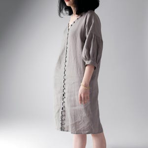 May include: A knee-length, light gray dress featuring a V-neckline and a vertical row of small, round buttons running down the entire front center. The garment has three-quarter length puff sleeves with gathered cuffs and a relaxed, straight fit.