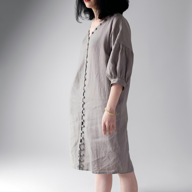 May include: A knee-length, light gray dress featuring a V-neckline and a vertical row of small, round buttons running down the entire front center. The garment has three-quarter length puff sleeves with gathered cuffs and a relaxed, straight fit.