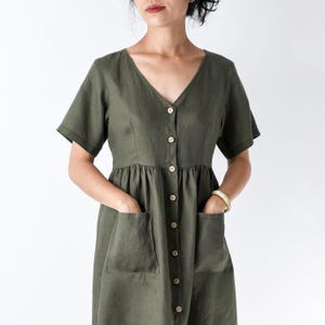 Peut inclure: Une robe vert olive dotée d'un col en V, de manches courtes et d'une fermeture boutonnée sur le devant avec des boutons clairs. Le vêtement comprend deux grandes poches plaquées sur le devant et une taille froncée, créant une silhouette décontractée.