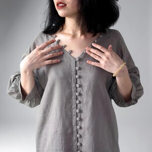 May include: A grey long-sleeved shirt featuring a V-neckline and a vertical row of matching fabric-covered buttons running down the center front. The garment has gathered sleeves and a textured appearance, with a simple, clean design.