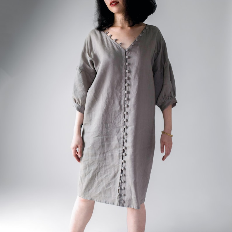 May include: A knee-length, light gray dress featuring a V-neckline and a vertical row of matching fabric-covered buttons running down the center front. The garment has three-quarter length sleeves with gathered cuffs and a relaxed, loose fit.