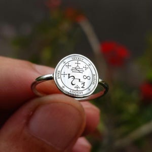 May include: A silver-toned ring featuring a circular face engraved with intricate symbols and the text Azrael, Xhay, Morth, and Adonay. The band is thin and polished, with a smooth finish that reflects light across the detailed circular emblem.