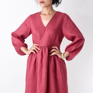 May include: A solid dark rose-colored dress featuring a V-neckline, long sleeves with gathered elasticized cuffs, and a fitted bodice that transitions into a slightly pleated skirt. The garment is constructed from a textured fabric.