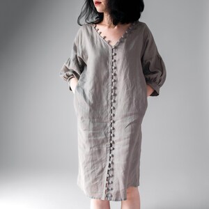 May include: A taupe-colored midi dress featuring a V-neckline, three-quarter length puff sleeves, and a full-length front button placket with matching round buttons. The garment has a loose, relaxed fit and includes side pockets for functionality.