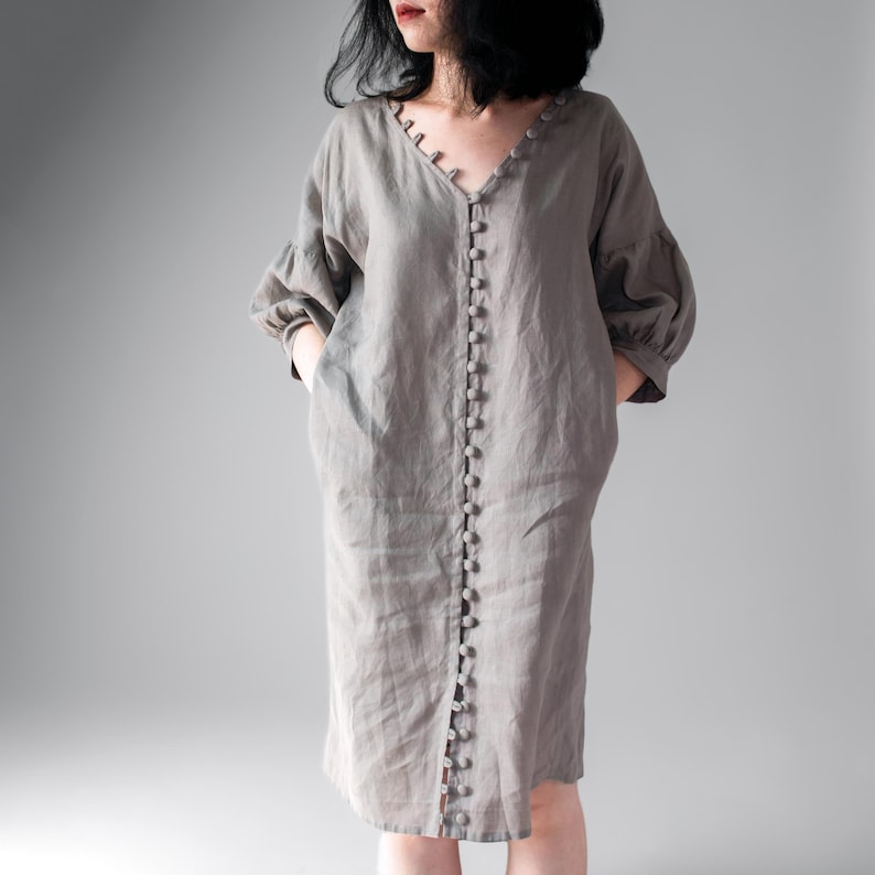 May include: A taupe-colored midi dress featuring a V-neckline, three-quarter length puff sleeves, and a full-length front button placket with matching round buttons. The garment has a loose, relaxed fit and includes side pockets for functionality.