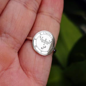 May include: A silver ring with a circular face featuring engraved symbols and text. The ring's face has a detailed design with a central symbol and surrounding text. The ring is held by a person.