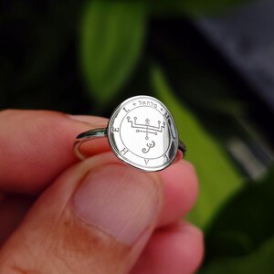 May include: A silver ring with an oval-shaped face featuring an engraved design with symbols and text. The ring is held against a blurred green background. The ring's band is thin and polished.