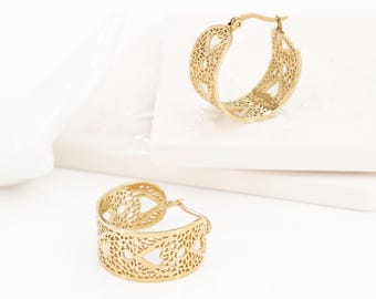 May include: A pair of gold-toned hoop earrings featuring a wide band with an intricate, laser-cut filigree pattern. The design incorporates repeating heart-shaped cutouts throughout the metal surface, finished with a hinged post closure.
