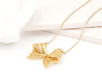 Puede incluir: Un collar de tono dorado con un delicado colgante de lazo de filigrana. El colgante está elaborado con intrincados patrones de alambre entrelazado que forman una figura de lazo tridimensional, suspendido de una fina cadena de metal dorado.