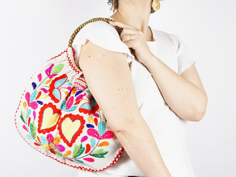 May include: A white fabric tote bag with a colourful embroidered design featuring hearts, flowers, and leaves. The embroidery is done in shades of red, pink, yellow, blue, and green.