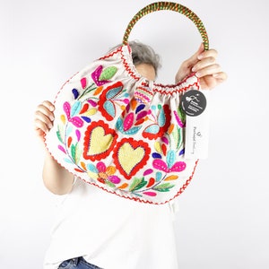 May include: White fabric tote bag with colourful embroidered flowers and hearts. The bag has a brown and green rope handle. The bag has a tag that says "Portugal Embroidery".
