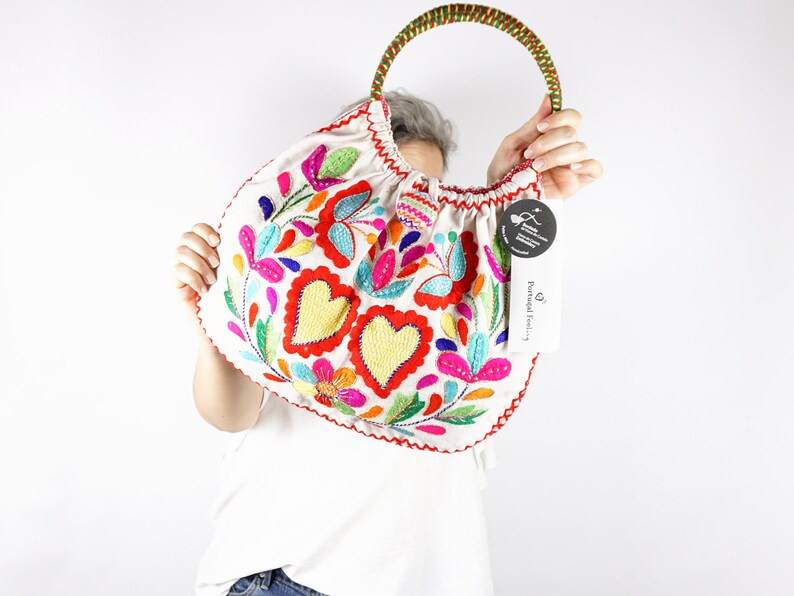 May include: White fabric tote bag with colourful embroidered flowers and hearts. The bag has a brown and green rope handle. The bag has a tag that says "Portugal Embroidery".