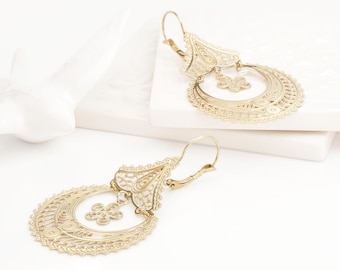 May include: A pair of gold-toned dangle earrings featuring intricate filigree metalwork. Each earring consists of a triangular top piece connected to a large circular hoop, with a small flower-shaped charm suspended in the center of the hoop.