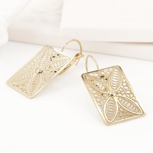 Pode incluir: Um par de brincos de filigrana em tom dourado com um delicado design floral. Os brincos são de forma retangular e têm um fecho de gancho.