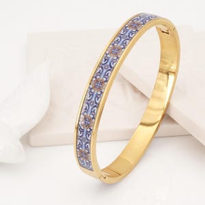 May include: A gold-toned hinged bangle bracelet featuring a central inlay of blue, white, and orange floral tile patterns. The metal surface is polished and reflective, with a secure clasp mechanism visible on the side of the circular band.