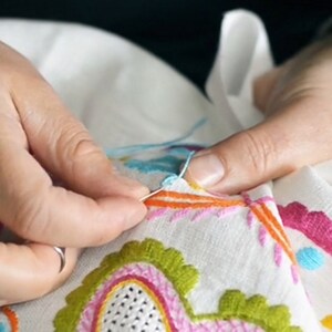 May include: Close-up of a person's hands embroidering a white fabric with colourful floral designs. The embroidery includes pink, green, orange, and blue thread.