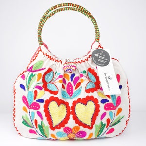 May include: White fabric tote bag with colourful embroidered flowers and hearts. The bag has a green and red handle and a small tag that says "Portugal Feeling".
