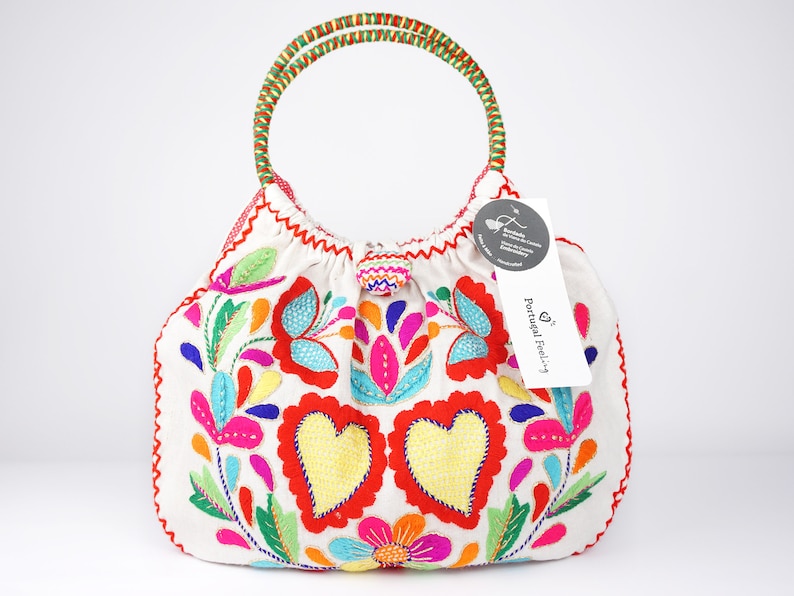 May include: White fabric tote bag with colourful embroidered flowers and hearts. The bag has a green and red handle and a small tag that says "Portugal Feeling".