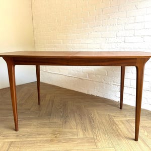 May include: A rectangular wooden dining table with a smooth, warm-toned finish. The table features four tapered legs that extend outward from the corners, supporting a flat surface with a visible seam indicating an extendable design feature.
