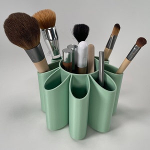 May include: A light green, flower-shaped makeup brush holder with eight slots. The holder is filled with various makeup brushes, including a large, fluffy brush, a smaller, angled brush, and a few thin, pointed brushes.