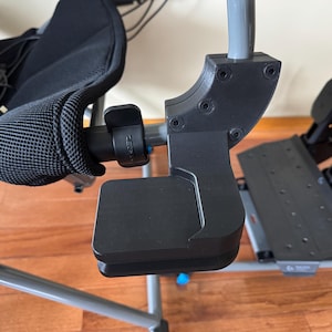 Peut inclure: Un fauteuil de jeu noir avec un dossier en maille et un repose-pieds. La chaise a un cadre gris et est conçue pour être utilisée avec un volant et des pédales de course. Les pédales sont noires et montées sur une plateforme grise.
