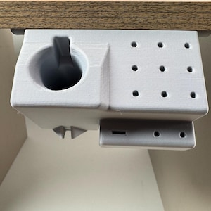 Peut inclure: Un organiseur de bureau gris clair, imprimé en 3D, conçu pour être fixé sous un bureau. Il comprend un porte-gobelet, une section avec neuf petits trous et une section plus petite avec deux trous. L'organisateur est monté sur un bureau en bois.
