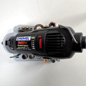 Peut inclure: Une perceuse rotative Dremel MultiPro noire à vitesse variable avec un contrôle de vitesse rouge. L'outil est monté dans un support gris avec divers accessoires. Le texte "Dremel MultiPro Variable Speed" est visible.