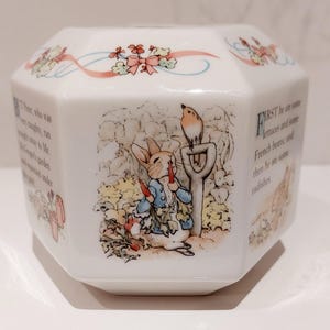 May include: A white octagonal ceramic container featuring illustrations of Peter Rabbit eating a carrot next to a garden spade and a small bird. The surface includes printed text from the story and a decorative border of pink ribbons and flowers.
