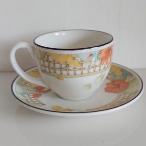 May include: A white teacup and saucer set with a floral pattern. The cup has a curved handle and a black rim. The saucer and cup feature orange, yellow, and green floral patterns with a trellis design.
