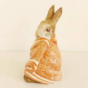 May include: A ceramic rabbit figurine, wearing a brown jacket with white stripes and polka dots. The rabbit has brown ears and a light brown face with red eyes. A decorative item.