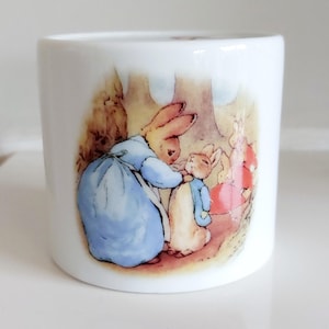 May include: White ceramic mug with a colourful illustration of two rabbits in a woodland scene. One rabbit is in a blue dress, the other in a blue jacket. The design is reminiscent of a children's storybook.